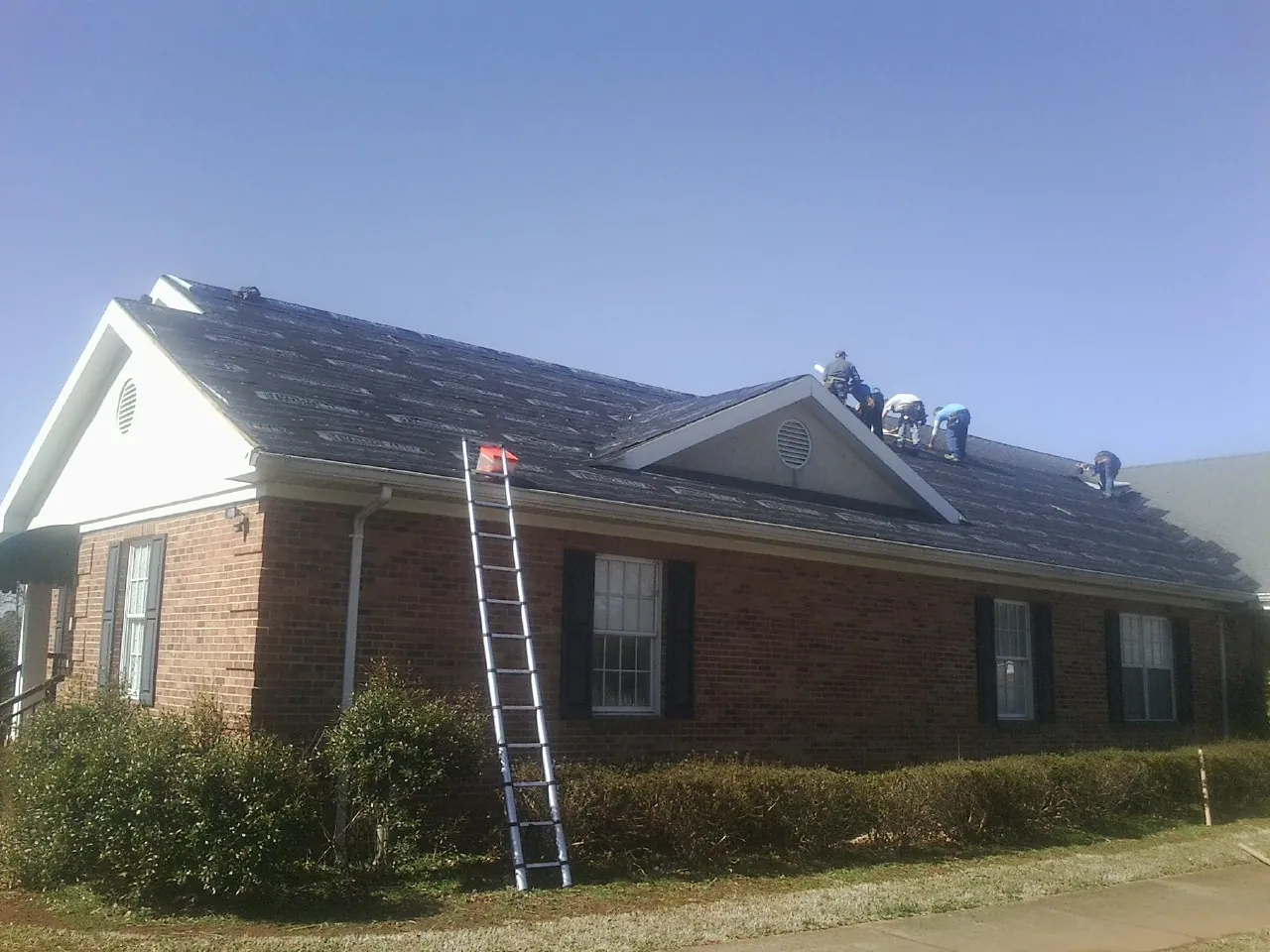 Crew tearing shingles off a brick ranch roof with a ladder set — Emergency Roof Repair work by our crew in Trego